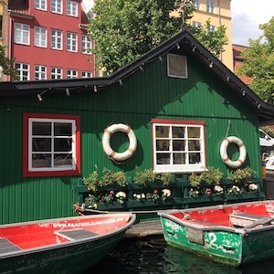 A green houseboat floats on a canal. Life preservers decorate its walls. Two smaller boats float in front of it.