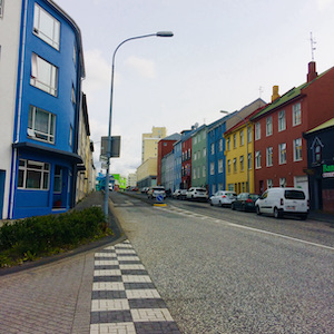 Buildings in primary colors line a city street. A checkerboard pattern marks a crosswalk in the foreground.