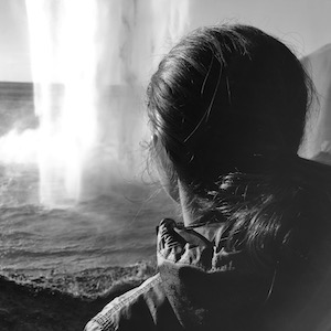 Shea Soler stands on a path behind a large waterfall and watches it.