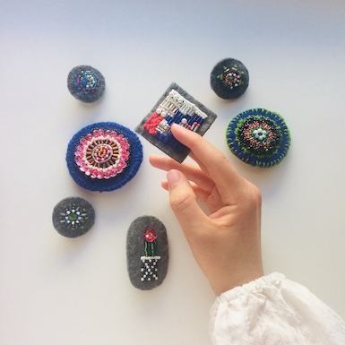 A hand holding a hand-embroidered, beaded brooch. Several other brooches of various sizes shown in the background.