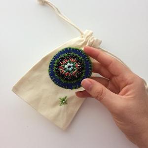 Blue felt circle embroidered in a radial pattern that resembles grass, black pebbles and pink wildflowers.