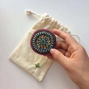 Gray felt circle embroidered with a radial pattern using little pearls, green sequins and glass beads in teal and pink.