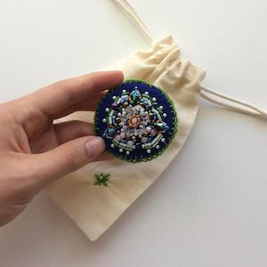 Blue felt circle embroidered with a flower using pearls and glass beads in green, white, blush, orange and iridescent blue.