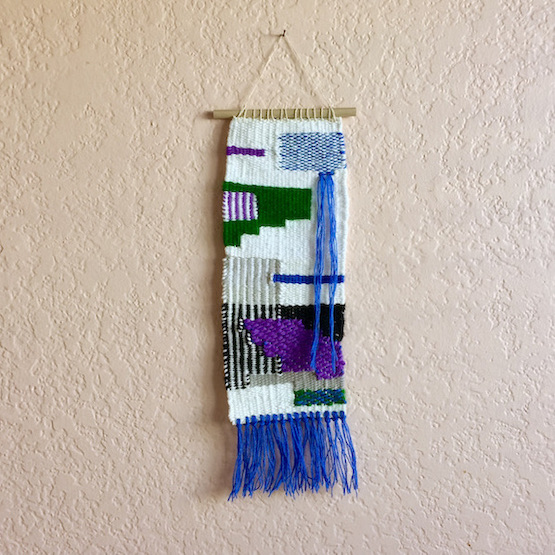 Rectangular weaving inspired by rock formations, featuring stripes, varigation, and overlapping shapes. In blue, purple, dark green, gray, white and black.